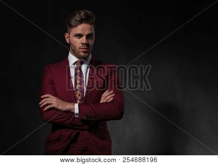 portrait of elegant man in grena suit standing with arms crossed and looking to side while standing on grey wallpaper background