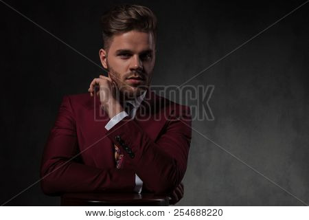 portrait of seductive man in gren suit leaning on a wooden round table while standing on grey wallpaper background