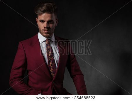 portrait of handsome elegant man wearing a red suit sitting on grey wallpaper background and looking to side