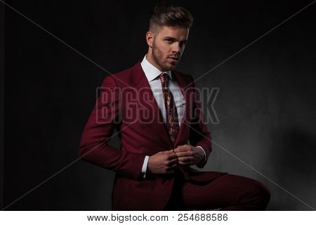 elegant man buttoning his red suit and sitting on a wooden chair on grey wallpaper background