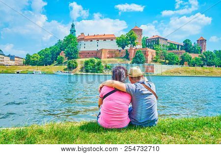 The Young Couple Sit On The Bank Of Vistule River Overlooking The Wawel Castle, Krakow, Poland