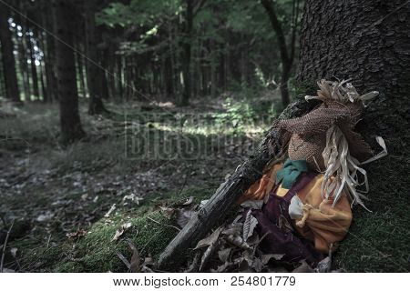 Scarecrow Sitting At The Roots Of A Tree, Holding A Branch, Covered With Dried Leaves, In A Dark For