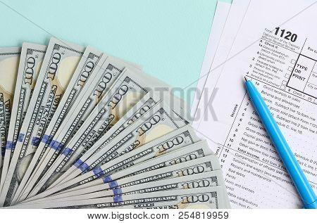 1120 Tax Form Lies Near Hundred Dollar Bills And Blue Pen On A Light Blue Background. Us Corporation