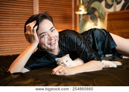 Beautiful chinese young woman wearing pajamas lies in bed. Lazy morning. Happy sleppy girl playing with her little kitty in bedroom.