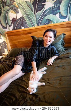 Beautiful chinese young woman wearing pajamas lies in bed. Lazy morning. Happy sleppy girl playing with her little kitty in bedroom.