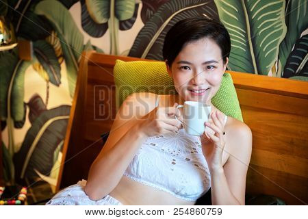 Beautiful chinese young woman wearing pajamas lies in bed and drinks coffee. Lazy morning. Happy sleppy girl wit cup og tea in bedroom.