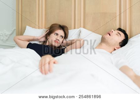 Annoyed Asian Wife Blocking Her Ears From Noise Of Husband Snoring In Bedroom At Home. Young Couple 