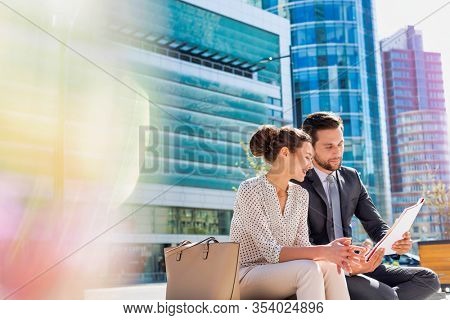 Young attractive businessman discussing business plans over document with beautiful businesswoman while sitting on bench against office building