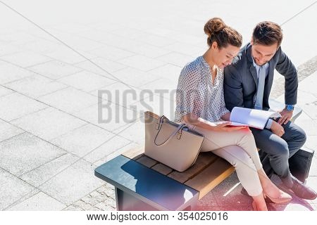 Young attractive businessman discussing business plans over document with beautiful businesswoman while sitting on bench against office building