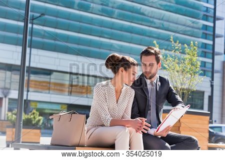 Young attractive businessman discussing business plans over document with beautiful businesswoman while sitting on bench against office building