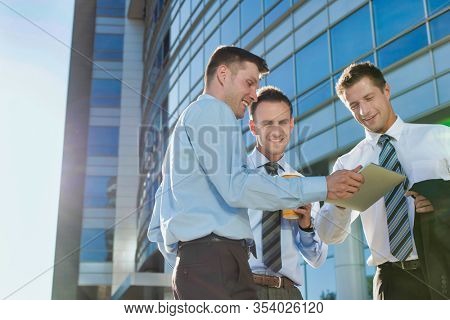 Mature attractive businessman showing and discussing plans over digital tablet with colleagues against office building