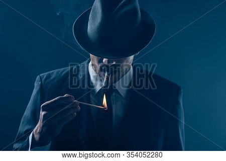 Mafioso With Covered Eyes With Felt Hat Lighting Cigar On Dark Blue Background