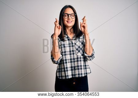 Young brunette woman with blue eyes wearing casual shirt and glasses over white background gesturing finger crossed smiling with hope and eyes closed. Luck and superstitious concept.