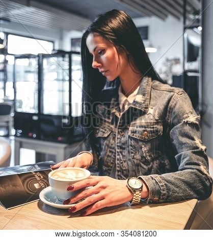 Pretty Face Brunette Girl With Cappucinno On Table Reading Magazine In Cafe