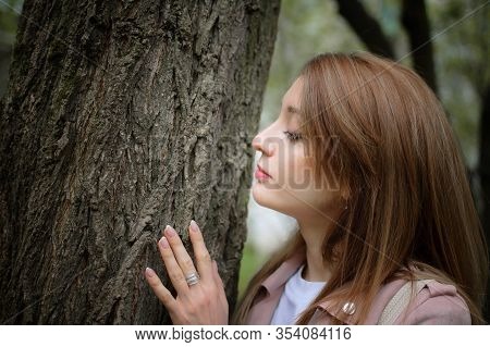 A Young Attractive Woman With A Blonde Hair Posing Near A Big Tree In A Spring Park Outdoors. She Le