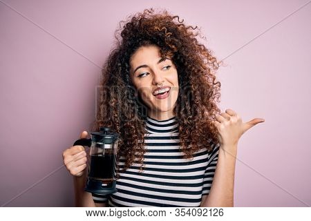 Young beautiful woman with curly hair and piercing doing coffee holding french coffeemaker pointing and showing with thumb up to the side with happy face smiling