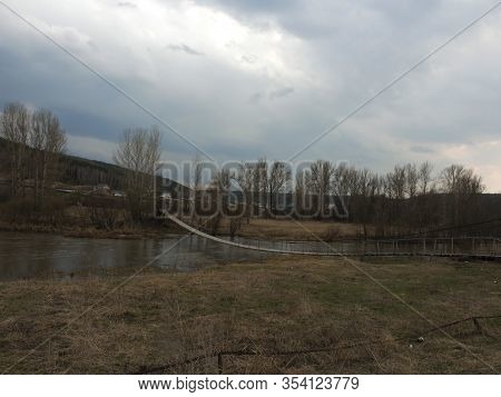 Trees, Dry Grass And A Wooden Rural Bridge Over A Small River. Rustic Old Bridge Over The River