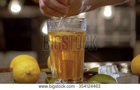 Close Up Male Hand Squeezing Lemon Into A Glass With Ice Tea Or Cocktail. Glass With A Refreshing Dr