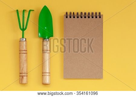 Wooden Handled Garden Hand Trowel And Hand Fork And Empty Noteboook On Yellow Background