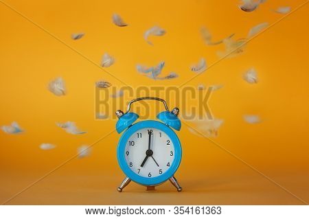 Soft Fluffy Flying Feathers And Blue Alarm Clock On Yellow Background. Wake Up Concept.