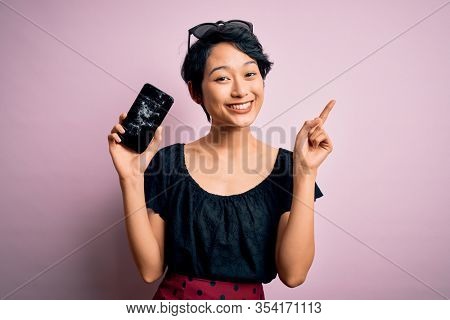 Young beautiful chinese woman holding broken smartphone showing craked screen surprised with an idea or question pointing finger with happy face, number one