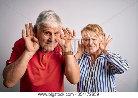 Senior beautiful couple standing together over isolated white background Trying to hear both hands on ear gesture, curious for gossip. Hearing problem, deaf