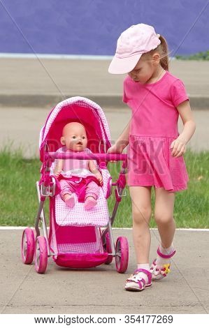 Caucasian Girl In A Pink Dress, Cap Rolls A Doll In A Pink Stroller. Children Games On The Street. F