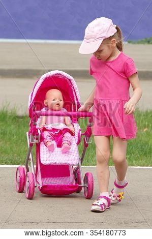 Caucasian Girl In A Pink Dress, Cap Rolls A Doll In A Pink Stroller. Children Games On The Street. F