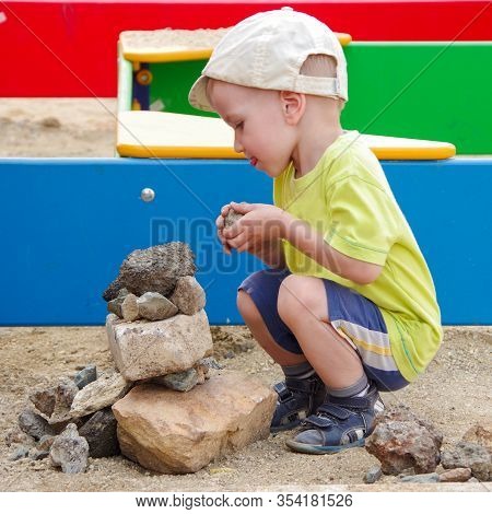 The Kid In A Yellow T-shirt, Cap, Dark Shorts Builds A Fortress Of Stones On A Platform On The Stree