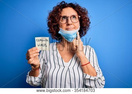 Middle age curly hair woman wearing medical mask holding reminder with corona virus message serious face thinking about question, very confused idea