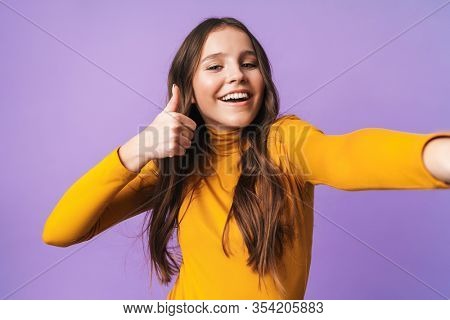 Image of young beautiful woman with long brown hair gesturing and taking selfie photo cellhone isolated over violet background