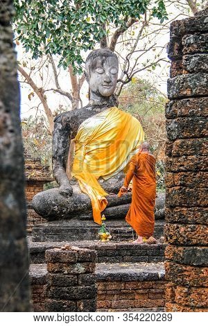 A Buddhistic Monk In Orange Dress Is Praying In Front Af An Ancient Statue