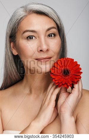 Beauty portrait of an attractive sensual mature topless woman with long gray hair standing isolated over gray background, posing with herbera flower