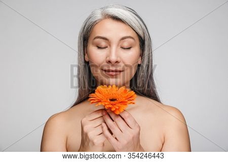 Beauty portrait of an attractive sensual mature topless woman with long gray hair standing isolated over gray background, ees closed