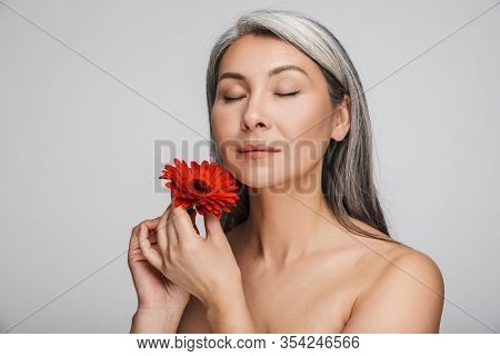 Beauty portrait of an attractive sensual mature topless woman with long gray hair standing isolated over gray background, posing with herbera flower