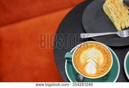 Cup Of Cappuccino With Latte Art On Black Metal Table.with Peacce Of Cake.against Orange Background.