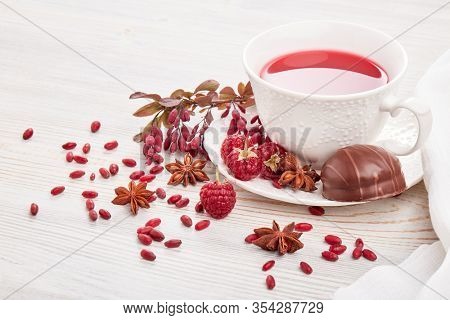 Healthy fruit tea with anise, barberries and raspberries in white porcelain cup and branch of berberry on light wooden background.