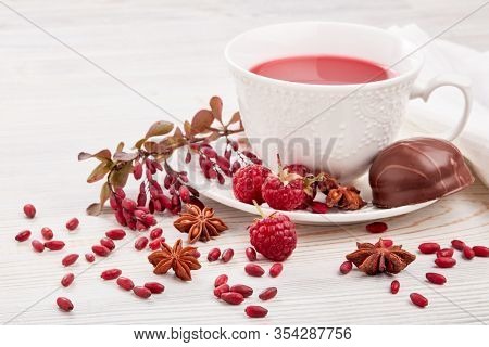 Healthy fruit tea with anise, barberries and raspberries in white porcelain cup and branch of berberry on light wooden background.