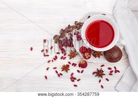 Healthy fruit tea with anise, barberries and raspberries in white porcelain cup and branch of berberry on light wooden background. Top view
