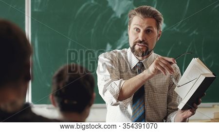 Vigorous Teacher Explaining Subject To Students Holding Open Book Pointing At It With Glasses Frame