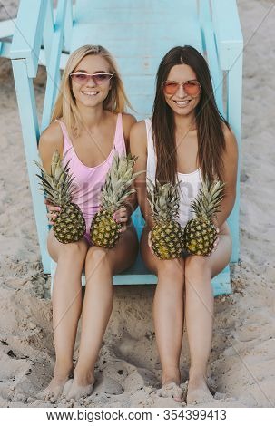 Two Cheerful Young Women Friends In Stylish Swimsuits And Sunglasses Holding Pinepples In Hands And 