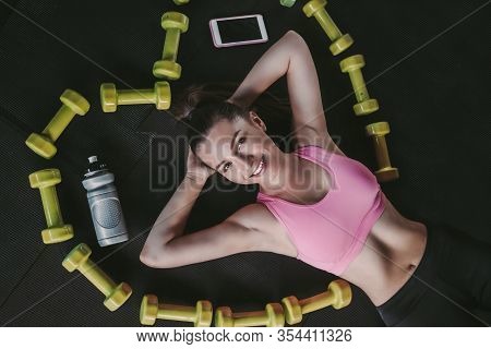 Top View Of Beautiful And Happy Athletic Girl In Sportswear Lying On Dark Floor And Smiling, While R