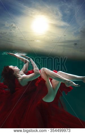 Mystical Underwater Portrait Of A Beautiful Young Woman In A Red Dress. The Girl Swims Under The Sur