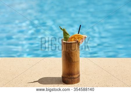 Outdoor Picture Of Wooden Cup With Cocktail Being Near Swimming Pool, Green And Piece Of Orange Bein