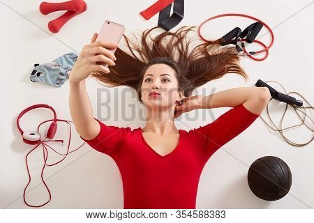 Woman Wearing Red Shirt Lying On White Surfaceholds Surrounded Different Sport Equipments, Expander,