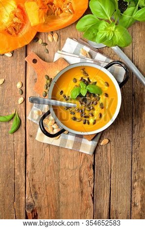 Enamel Pot Of Cream Pumpkin, Carrot Soup With Fresh Basil, Pumpkin Seed, Knife And Laddle On Rustic 