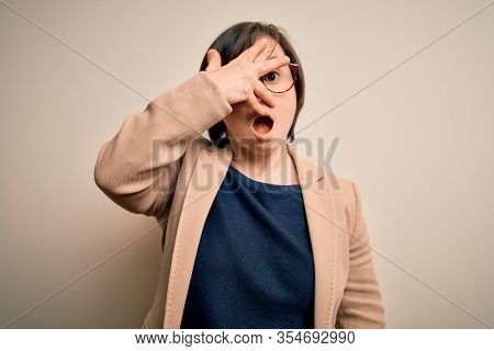 Young down syndrome business woman wearing glasses standing over isolated background peeking in shock covering face and eyes with hand, looking through fingers with embarrassed expression.