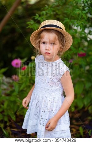 Sad Little Girl In A Hat And White Dress Among The Greenery In Summer
