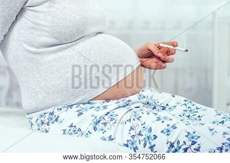 Pregnant Woman With A Cigarette In Her Hands, Sitting On The Bed. Hands With A Cigarette And Stomach