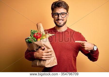Young man wearing glasses holding paper bag with food over isolated yellow background with surprise face pointing finger to himself